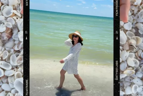 Black film-frame overlay taped onto a bed of small white shells, featuring a smiling woman in a straw hat and white crochet cover-up wading along the shoreline.